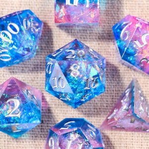 May include: A set of seven polyhedral dice featuring a translucent blue and pink gradient with embedded iridescent foil flakes. Each die has silver-colored numbers engraved on its faces, including a d20, d12, d10, d8, d6, d4, and a percentile die.