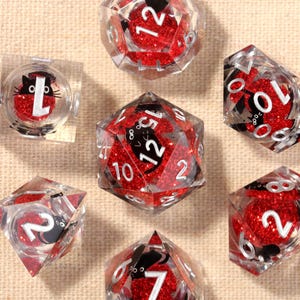 Può includere: Un set di sette dadi trasparenti a più facce con glitter rossi e numeri bianchi. Ogni dado presenta un'illustrazione di un gatto nero. I dadi sono disposti su una superficie beige.