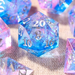 May include: A set of polyhedral dice featuring a translucent blue and pink gradient with embedded iridescent glitter flakes. Each die has sharp edges and clear, silver-colored engraved numbers. The collection includes various shapes such as a d20, d12, d10, d8, d6, and d4, all displaying a shimmering, multi-colored internal effect.