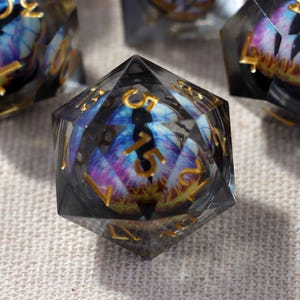 Puede incluir: Primer plano de varios dados poliédricos con un aspecto oscuro y ahumado. Cada dado presenta un diseño vibrante en forma de ojo con tonos de azul, morado y dorado. Los números son dorados. Los dados son probablemente para juegos de mesa.