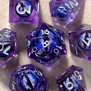 May include: A set of seven purple resin dice with a fantasy eye design. Each die features a different number in silver, with a detailed eye graphic in the center. The dice are arranged on a neutral-toned surface.