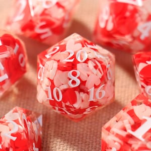 May include: A collection of polyhedral dice with a clear resin base, filled with red and pink heart-shaped confetti. The dice feature white numbering. The set includes various shapes, including a 20-sided die.
