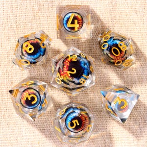 Puede incluir: Un juego de siete dados poliédricos hechos de resina transparente, cada uno con un ojo realista multicolor con iris azul y pupila de color naranja rojizo intenso. Los números están grabados en un acabado dorado metálico en las caras.