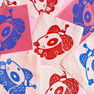 May include: A collection of fabric squares with hand-printed images of cartoon dogs wearing party hats. The dogs are printed in red and blue ink on a natural-colored fabric.