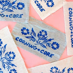 May include: Several fabric patches with a blue floral design and the text "CORNING*CORE" printed on them. The patches are in various shapes and sizes, with a pink background.