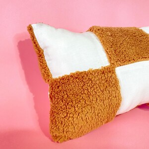 May include: A long rectangular pillow with a checkered pattern. The pillow is made of two different fabrics: a white cotton fabric and a brown fuzzy fabric. The pillow is lying on a pink surface.