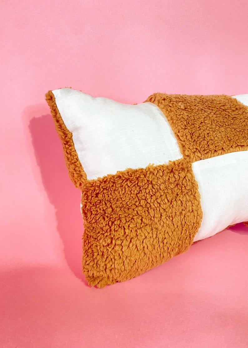 May include: A long rectangular pillow with a checkered pattern. The pillow is made of two different fabrics: a white cotton fabric and a brown fuzzy fabric. The pillow is lying on a pink surface.