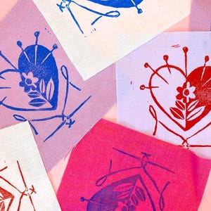 May include: A collection of fabric squares with heart-shaped floral designs. The hearts are printed in blue and red ink, with floral and pin details. The fabric colors include pink, lavender, and white, arranged on a pink background.