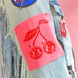 May include: A denim jacket with a pink patch featuring two red cherries with a disco ball design. The cherries have a green stem and leaves. The jacket has a blue and white "BOWIE" patch on the front.