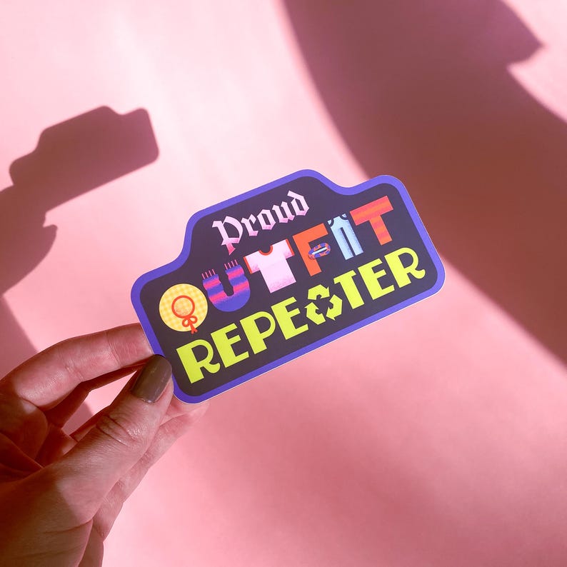 May include: A sticker with the text "Proud Outfit Repeater" in a stylized font. The sticker features a black background with colorful illustrations of clothing items and a recycling symbol. The sticker is held by a hand against a pink background.