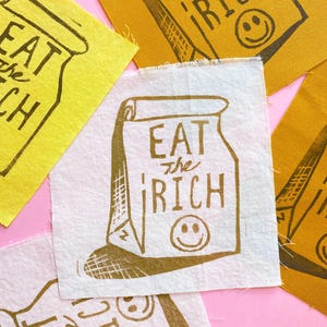 May include: Four fabric squares with the phrase "Eat the Rich" printed in brown on each. The squares are in different colors: yellow, white, and a darker yellow. Each square features a cartoon drawing of a paper bag with a smiley face.