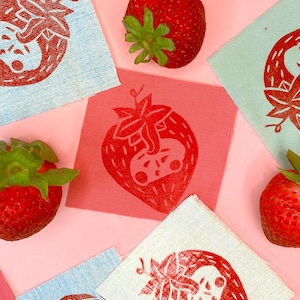 May include: A collection of red strawberry prints on various fabric squares, including light blue, pink, and white. Fresh strawberries are scattered around the fabric squares on a pink background. The strawberry prints have a unique skull design.