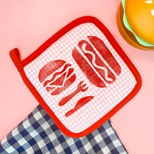 May include: A red-trimmed, square pot holder with a white and pink gingham pattern. The pot holder features red printed images of a hamburger, hot dog, fork, and knife. A blue and white checkered cloth is in the background.