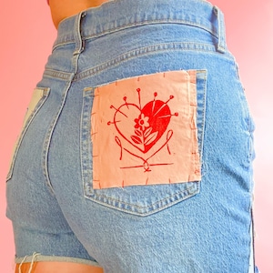 May include: Blue denim shorts with a pink patch on the back pocket. The patch features a red heart with a flower and pins.