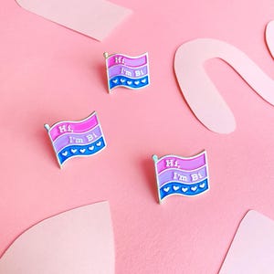 May include: Three enamel pins in the shape of a bisexual pride flag. Each pin says "Hi, I'm Bi" with a row of small hearts at the bottom.