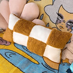 May include: A brown and white checkered throw pillow with a soft, fuzzy texture. The pillow is lying on a colorful patterned rug.