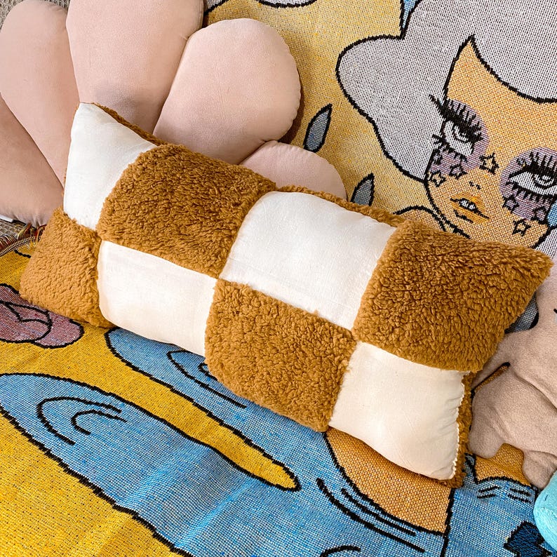 May include: A brown and white checkered throw pillow with a soft, fuzzy texture. The pillow is lying on a colorful patterned rug.