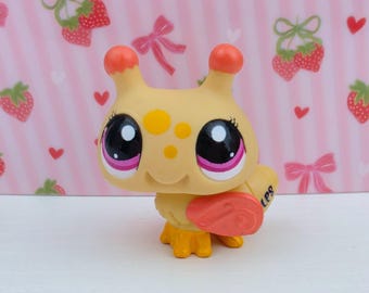 May include: A yellow and orange toy figure with large black eyes, pink accents, and a smiling face. The figure has orange antennae and a small orange object on its side. The background is pink with strawberry and bow patterns.