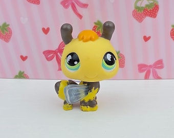 May include: A yellow and black toy bee with large, expressive eyes and a smiling face. The bee has brown antennae and a small orange tuft of hair. Its wings are a translucent blue-gray. The toy is a collectible figurine.