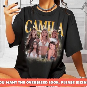 May include: Black oversized t-shirt featuring the name "CAMILA" in gold lettering with a vintage design. The shirt includes images of several women and the text "IF YOU WANT THE OVERSIZED LOOK, PLEASE SIZING UP".