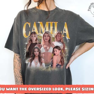 May include: Dark gray t-shirt featuring the name "CAMILA" in gold lettering above a collage of five women. The shirt has a vintage look, with the text "IF YOU WANT THE OVERSIZED LOOK, PLEASE SIZING UP" at the bottom. The PaperBloomArtStore logo is in the upper right corner.
