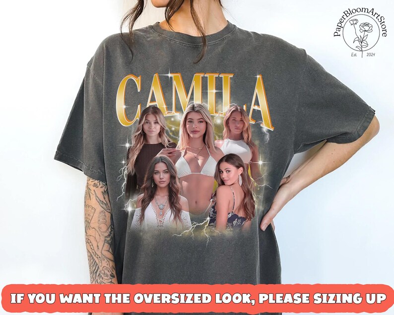May include: Dark gray t-shirt featuring the name "CAMILA" in gold lettering above a collage of five women. The shirt has a vintage look, with the text "IF YOU WANT THE OVERSIZED LOOK, PLEASE SIZING UP" at the bottom. The PaperBloomArtStore logo is in the upper right corner.