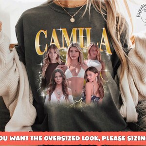 May include: Dark green sweatshirt featuring the name "CAMILA" in gold lettering with a vintage design. The sweatshirt has a graphic of five women and lightning bolts. The text at the bottom reads "IF YOU WANT THE OVERSIZED LOOK, PLEASE SIZING UP."