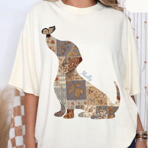 Puede incluir: Una camiseta de manga corta color marfil con el gráfico de un perro salchicha compuesto por varios patrones florales y geométricos en tonos marrón y beige, con una pequeña mariposa en la nariz y el nombre Molly impreso en letra azul.