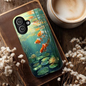 May include: A smartphone case featuring an underwater scene with orange and white koi fish swimming among green aquatic plants and lily pads. The case is resting on a brown leather-bound book, next to a cup of coffee.