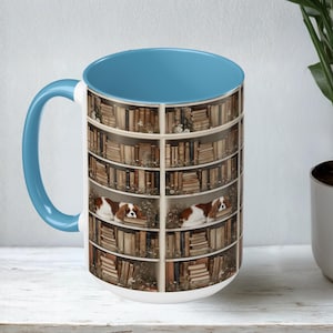 May include: A white ceramic mug with a blue interior and handle. The mug features a design of a bookshelf filled with books and two Cavalier King Charles Spaniels. The dogs are brown and white.