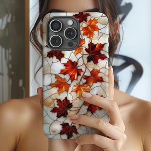 May include: A smartphone case featuring a stained-glass design with red, orange, and yellow maple leaves. The leaves are set against a white and cream background with dark brown lines, creating a mosaic effect. The case has a black border and a camera cutout.
