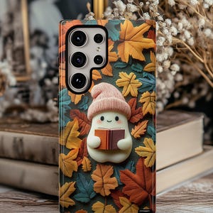 May include: A phone case featuring a whimsical design with a cartoon ghost wearing a pink hat and holding a book. The case is adorned with colorful autumn leaves in shades of orange, yellow, and green.