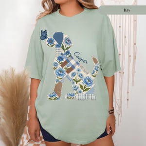 May include: A sage green short-sleeve t-shirt featuring a graphic of a dog silhouette filled with a blue and white floral and plaid patchwork pattern. A small blue butterfly sits on the dog's nose, and the name Cooper is printed in blue script.