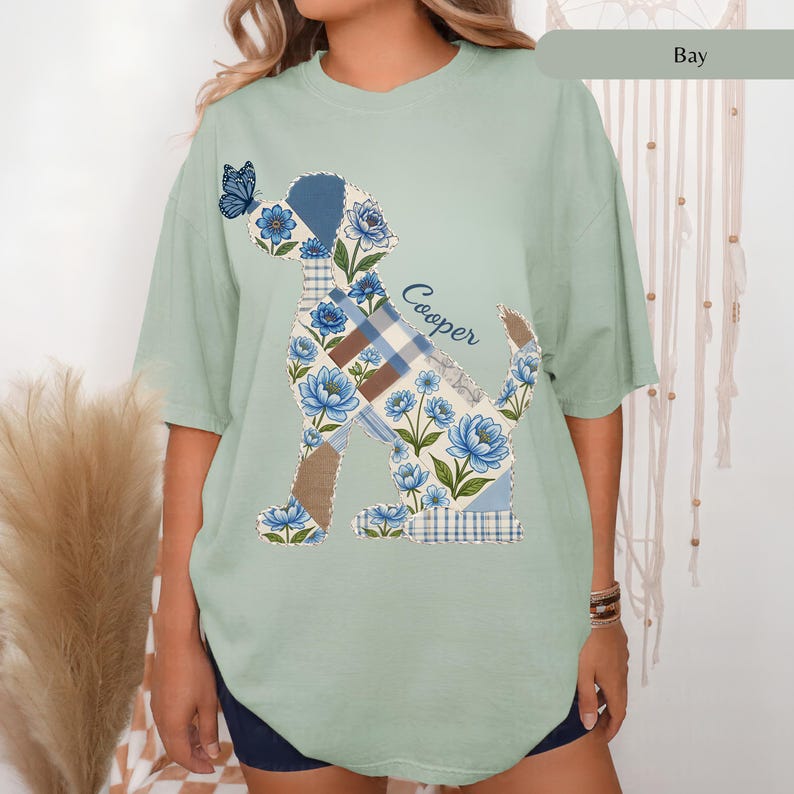 May include: A sage green short-sleeve t-shirt featuring a graphic of a dog silhouette filled with a blue and white floral and plaid patchwork pattern. A small blue butterfly sits on the dog's nose, and the name Cooper is printed in blue script.