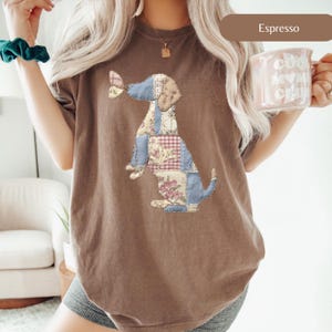 Może przedstawiać: Koszulka w kolorze espresso z patchworkowym wzorem psa. Pies składa się z różnych wzorów tkanin, w tym kwiatowych, w kratkę i gładkich niebieskich łatek. Koszulka ma luźny krój i krótkie rękawy. Słowo "Espresso" jest wydrukowane na brązowym pasku.