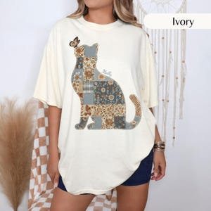 Puede incluir: Una camiseta de manga corta color marfil con un gran gráfico de un gato compuesto por varios patrones de patchwork florales y de cuadros en tonos azul, marrón y crema. Una pequeña mariposa descansa sobre la cabeza del gato y el nombre Molly está impreso.