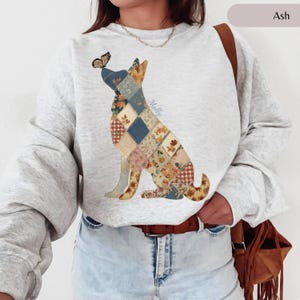 Puede incluir: Una sudadera de cuello redondo color gris ceniza con un gráfico de un perro sentado compuesto por varios patrones de tela de patchwork en tonos tierra. Una pequeña mariposa descansa sobre la nariz del perro y el nombre Molly está impreso en azul.