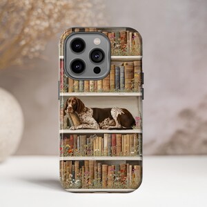 May include: A phone case featuring a detailed illustration of a dog resting on a bookshelf. The design includes rows of vintage books, flowers, and a brown and white dog. The case has a glossy finish.