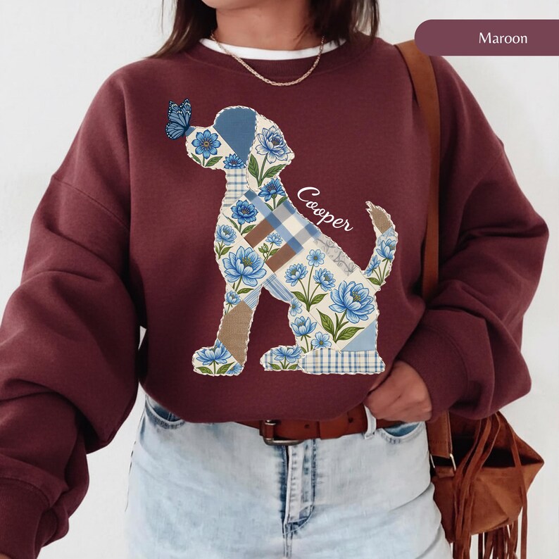 May include: A maroon crewneck sweatshirt featuring a large graphic of a dog silhouette filled with a blue floral and plaid patchwork pattern. A small blue butterfly rests on the dog's nose, and the name Cooper is written in white script.