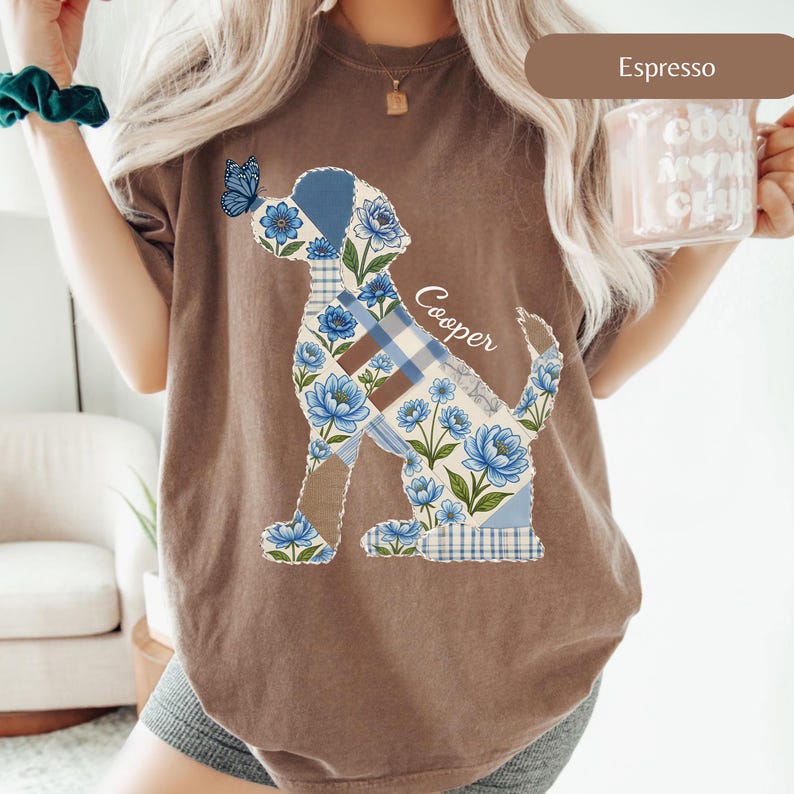 May include: An espresso brown short-sleeve t-shirt featuring a graphic of a dog silhouette filled with blue floral and plaid patterns. A blue butterfly rests on the dog's nose, and the name Cooper is written in white script next to the dog.