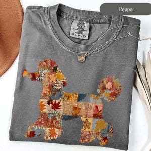 Op de afbeelding: Een grijs Comfort Colors T-shirt met een patchwork poedel ontwerp in herfstkleuren. Het shirt heeft een ronde hals en korte mouwen. Een zilveren ketting met een kleine hanger is over het shirt gedrapeerd. Het shirt is gelabeld "Pepper".