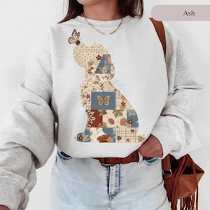 Peut inclure: Un sweat-shirt à col rond gris cendré arborant un grand motif représentant la silhouette d'un chien composée de divers motifs en patchwork dans des tons bleu, marron et crème, avec un papillon posé sur la tête et un autre sur le corps.