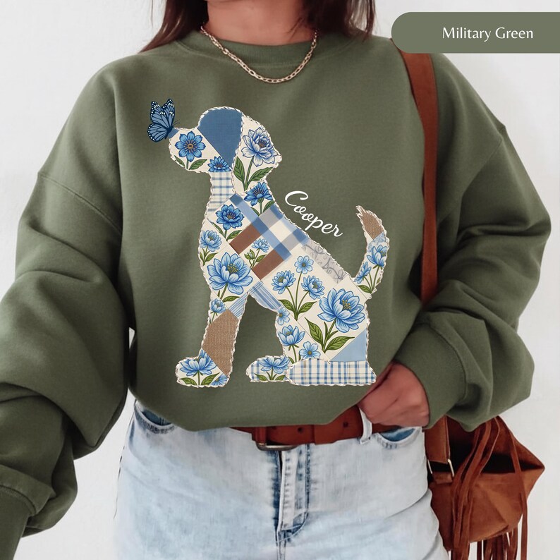 May include: A military green crewneck sweatshirt featuring a graphic of a dog silhouette composed of blue floral and plaid patchwork patterns. A blue butterfly rests on the dog's nose, and the name Cooper is written in white script text.