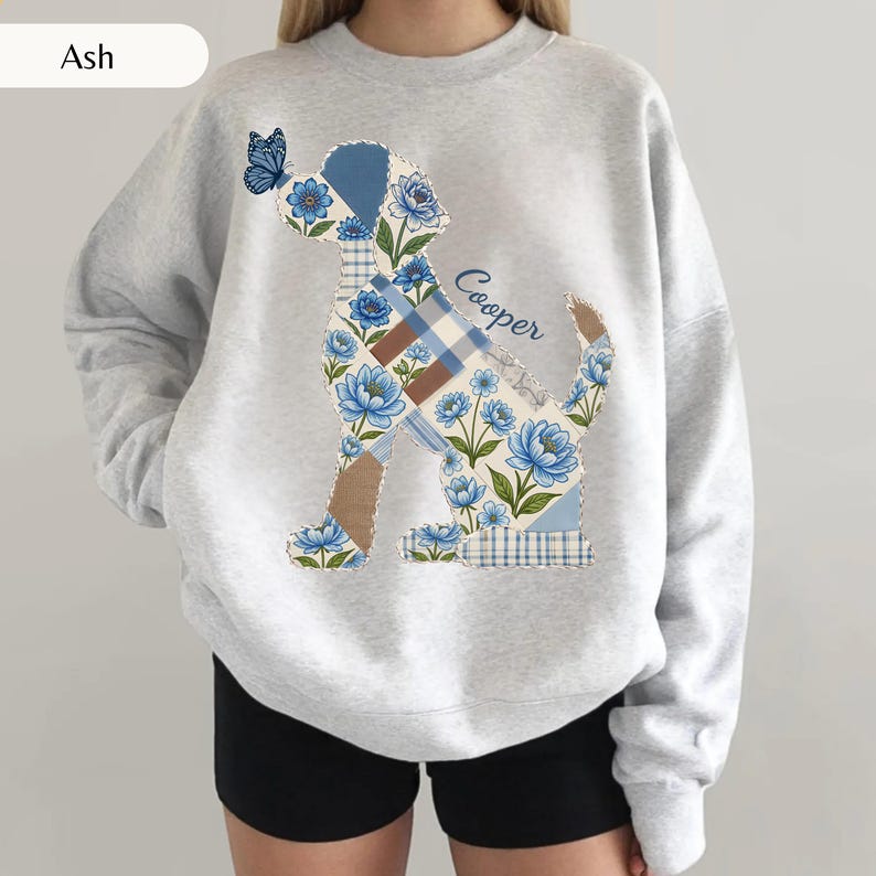 May include: An ash gray crewneck sweatshirt featuring a large graphic of a dog silhouette filled with a patchwork pattern of blue flowers, brown and blue plaid, and solid blue sections. A blue butterfly sits on the dog's head, and the name Cooper is written in blue script.