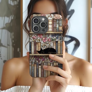 May include: A phone case featuring a bookshelf design with a sleeping black cat. The case has a floral and book pattern in shades of brown, pink, and blue. The phone case is held in front of a person.