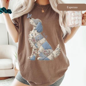 以下が含まれることがあります： エスプレッソカラーのTシャツ。花柄と青色のパッチワーク犬のデザイン。犬の鼻には蝶がいます。「Molly」の文字が犬の横に書かれています。このシャツは快適でカジュアルなスタイルです。