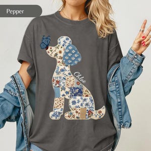 Puede incluir: Una camiseta gris carbón con un gráfico de un perro compuesto por varios parches con estampados florales en azul, blanco y beige. El perro mira a una mariposa azul posada en su nariz y el nombre Chloe está impreso en letra cursiva blanca.