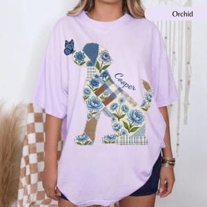 May include: A light purple short-sleeve t-shirt featuring a graphic of a dog composed of blue floral and plaid patchwork patterns. A blue butterfly rests on the dog's nose, and the name Cooper is printed in blue script next to the dog's back.