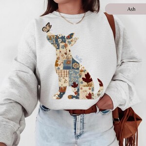 May include: An ash grey crewneck sweatshirt featuring a patchwork-style graphic of a sitting dog. The dog silhouette is composed of various floral and maple leaf patterns in shades of blue, brown, and cream, with a butterfly perched on its nose.