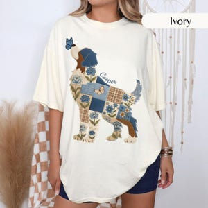 Puede incluir: Una camiseta de manga corta color marfil con un gran gráfico de un perro compuesto por patrones de patchwork en azul, marrón y crema con motivos florales y mariposas. El nombre Cooper está impreso en azul sobre el lomo del perro.